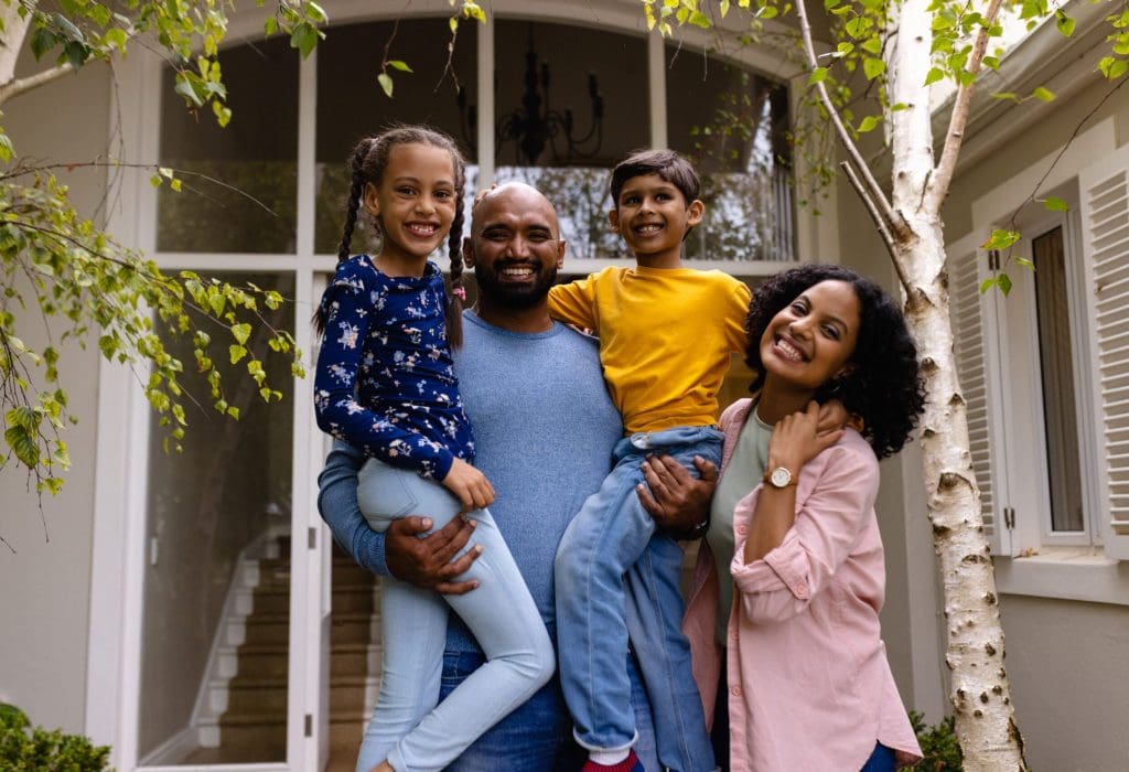 Portrait of happy biracial parents holding son and daughter, embracing in garden outside house. Family, love, togetherness, home, lifestyle and domestic life, unaltered. Portrait of happy biracial parents holding son and daughter, embracing in garden outside house. Family, love, togetherness, home, lifestyle and domestic life, unaltered.